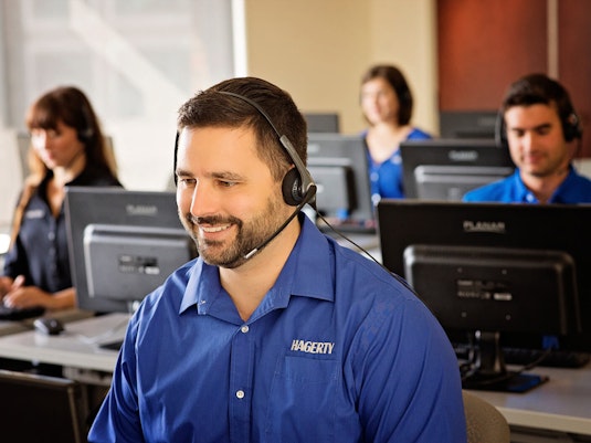 A man wearing a headset and a blue Hagerty uniform smiles widely. Behind him are other uniformed employees sitting and working at computers.
