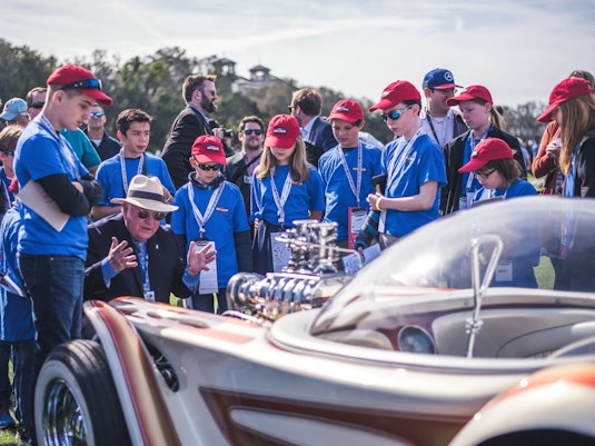 A sitting senior man speaks outside surrounded by a large group of attentive kids and teenagers, and gestures both hands toward a retrofuturistic car in front of him.