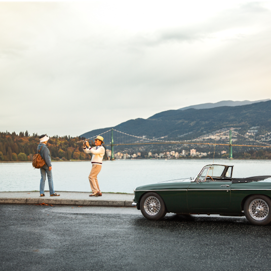 A man and woman stand outside a dark green collector convertible next to water with a bridge and mountains in the background.