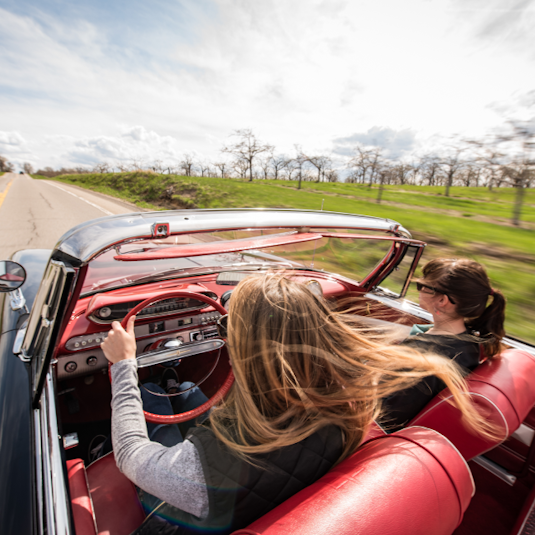 2 women driving a collector convertible down a country road.