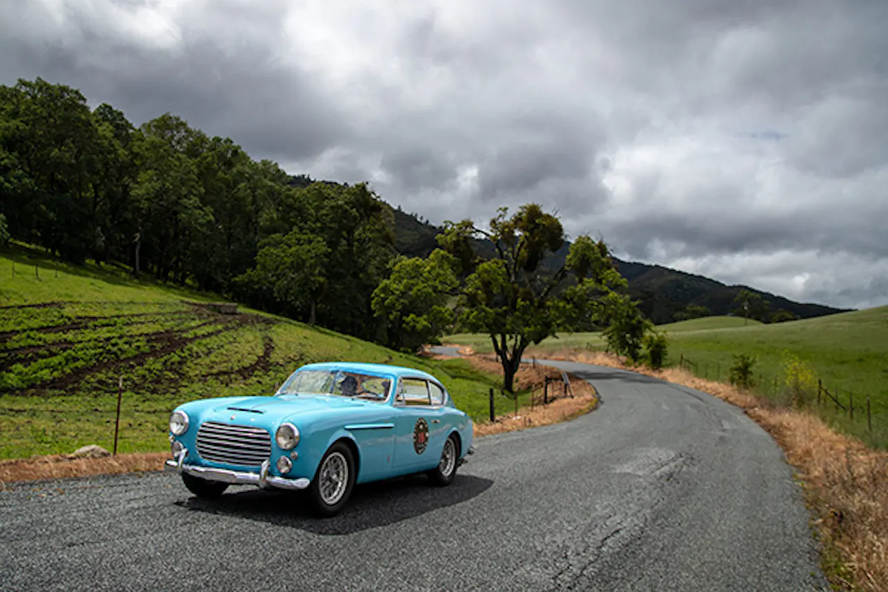A baby blue vintage collector vehicle driving on a winding country road on a cloudy day