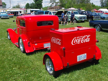 A red collector Coca-cola vehicle and trailer parked on grass in front of other collector vehicles.