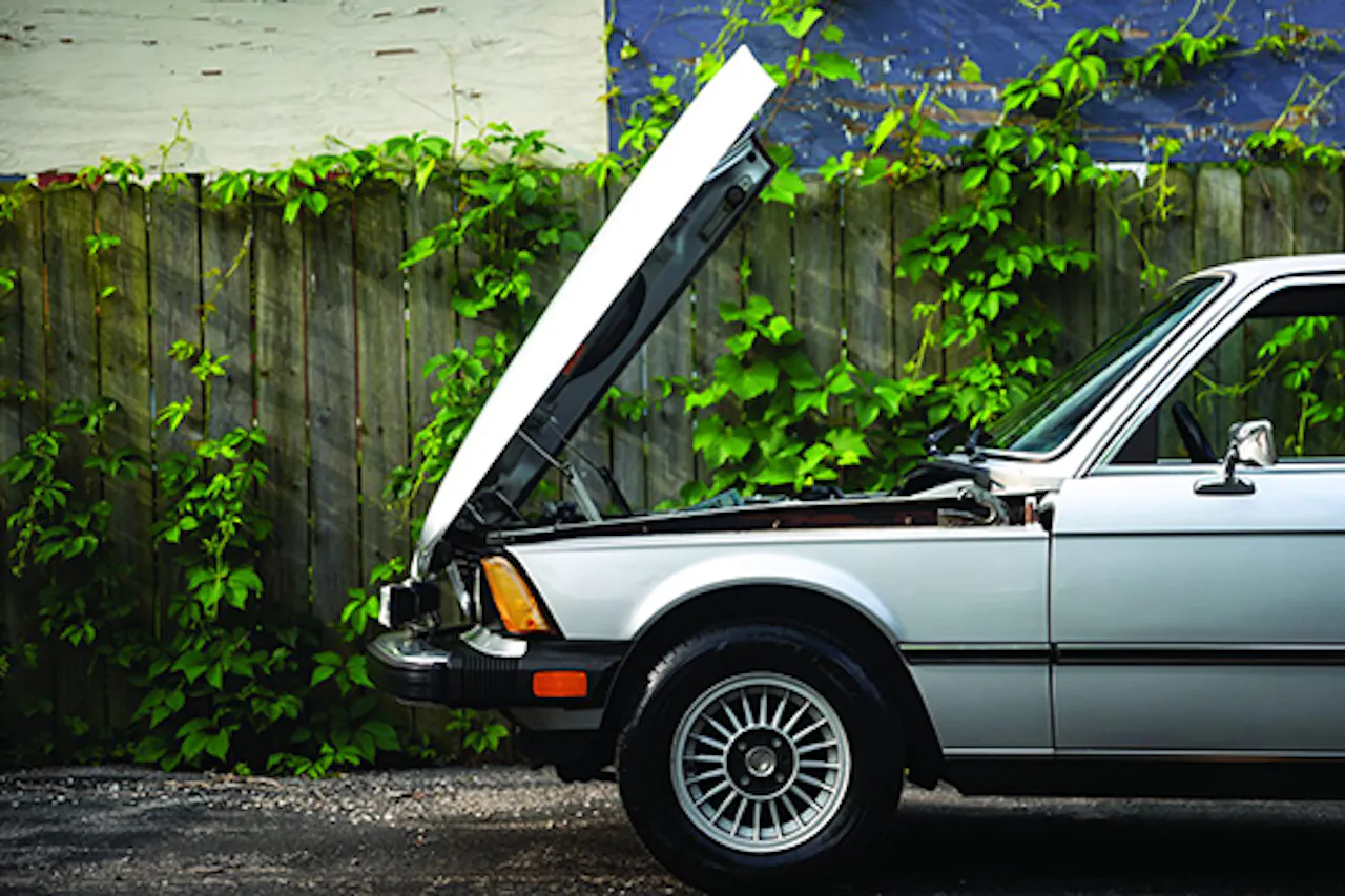 A silver classic car with it's hood raised open parked in front of a wooden fence covered in vines