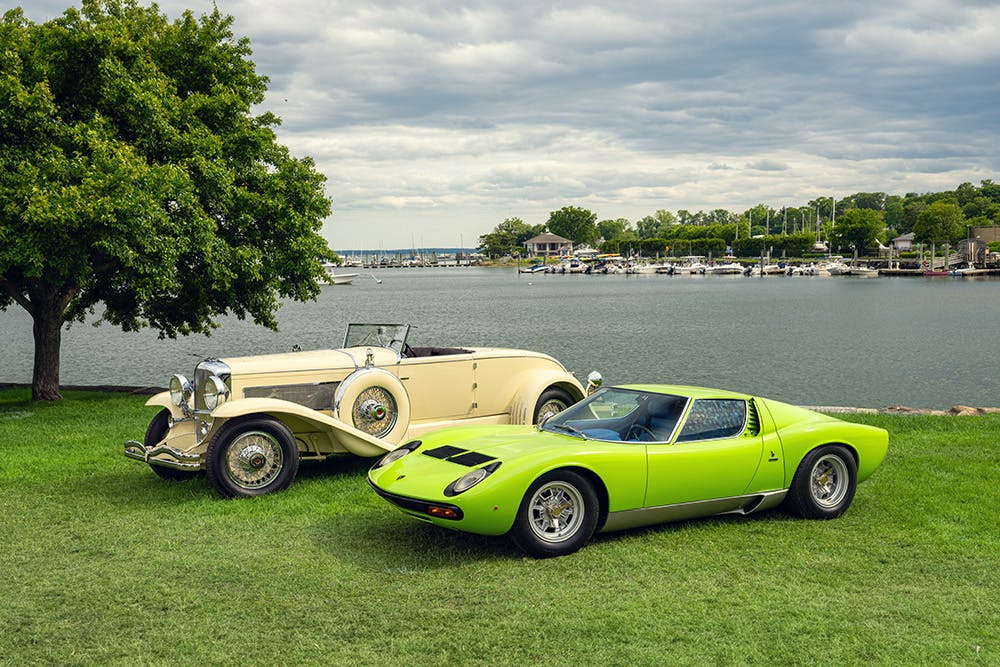 Two collector vehicles-one lime green and one off white-parked on the grass in front of a lake at the Greenwich Concours.