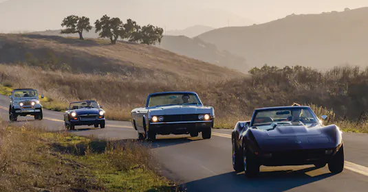 4 classic cars driving down the road in a line.