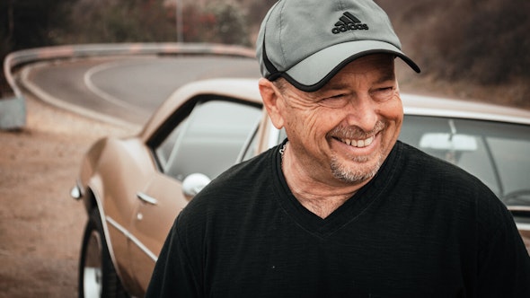 A man smiling off-camera, with a collector car and road in the background.