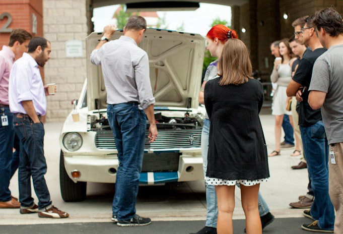 A group of collector car enthusiasts gather around a white Mustang with the hood popped up.