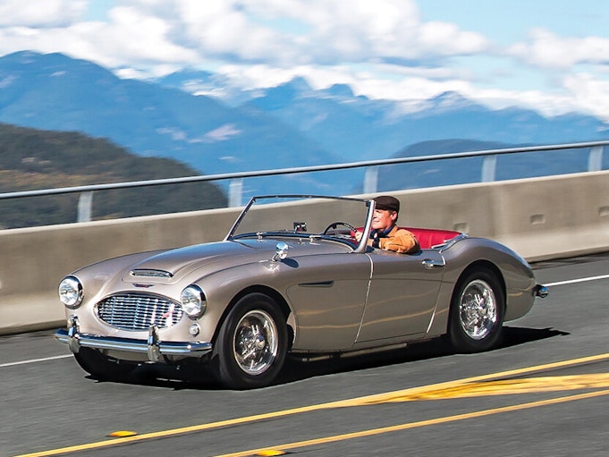 A man driving a collector car down a road, with mountains and blue sky in the background.