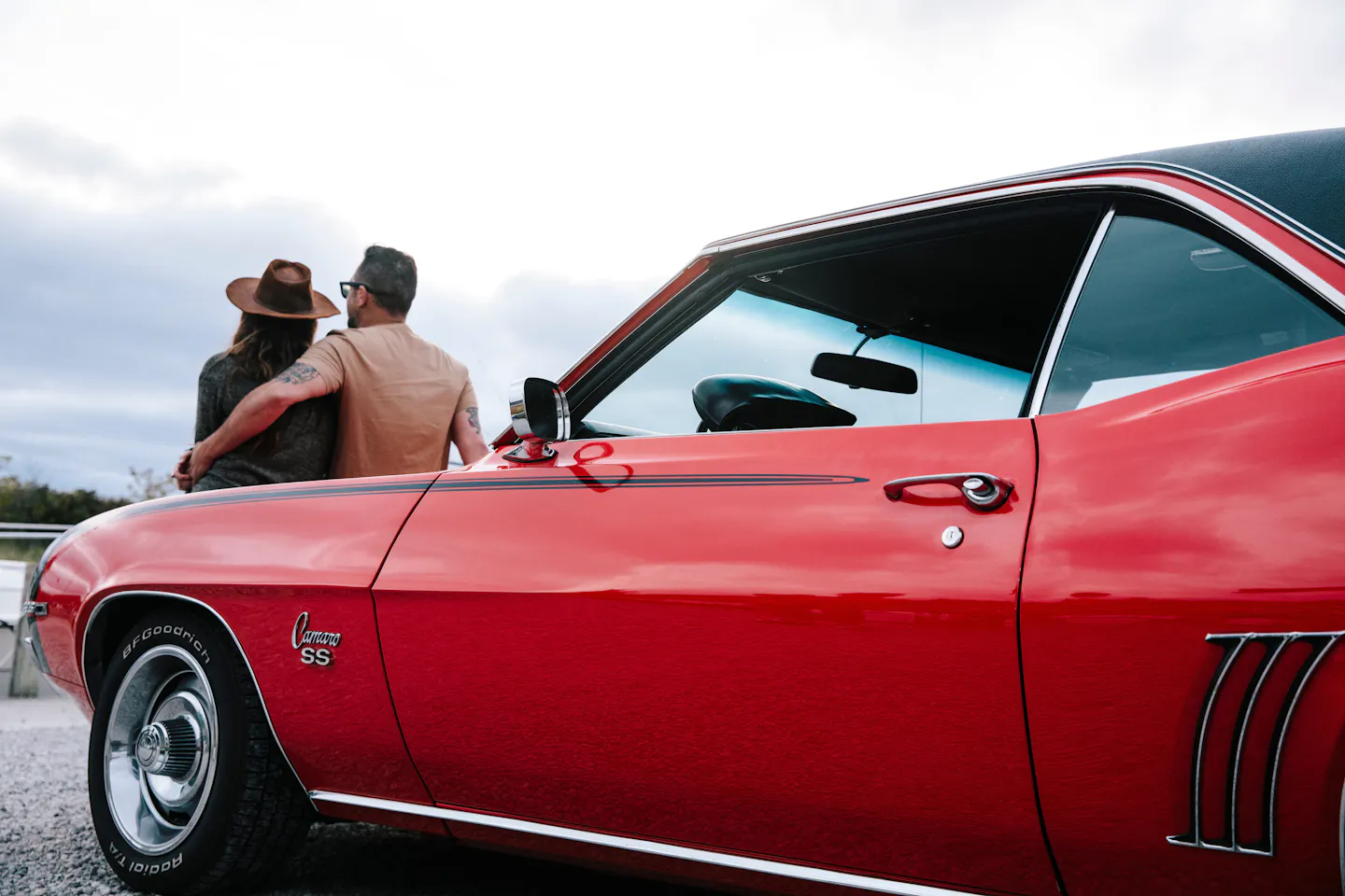 A couple sits on the hood of a red 1969 Camaro, looking out at the view