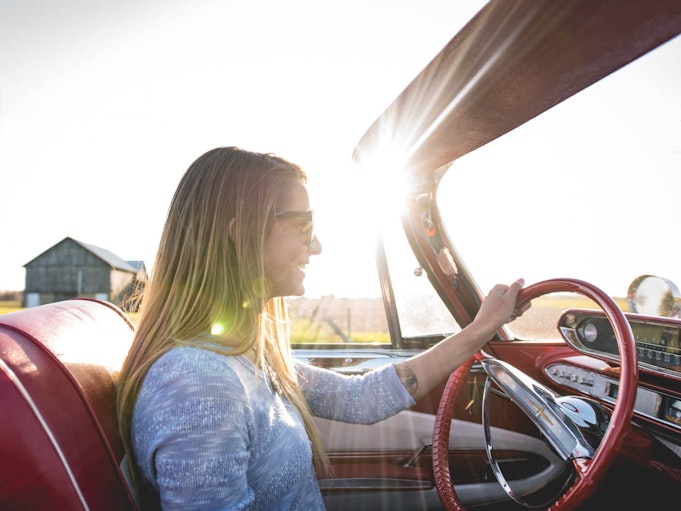 Bright sunshine beams down on a woman happily driving a red convertible.