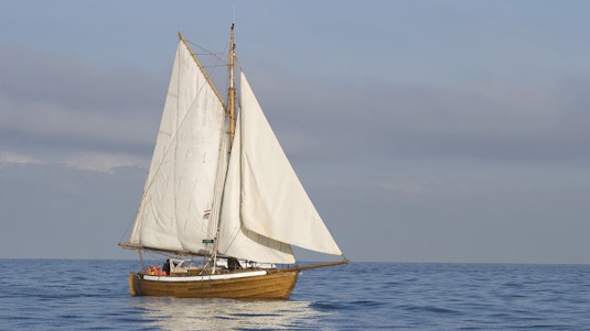 A wooden collector sailboat with white sails unfurled, with gray clouds in the background.