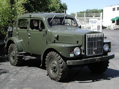 A dark green military vehicle in a parking lot, with bushes and a building in the background.