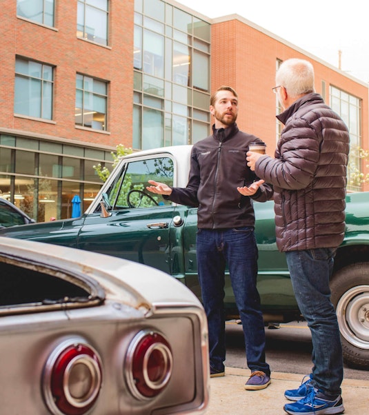 two men having a conversation next to two classic collector cars