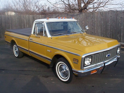 A yellow collector pickup truck parked on concrete in front of a wooden fence.