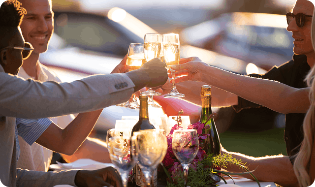 A group of four people toasting champagne in the sunset while sitting in front of a row of classic vehicles at a car show