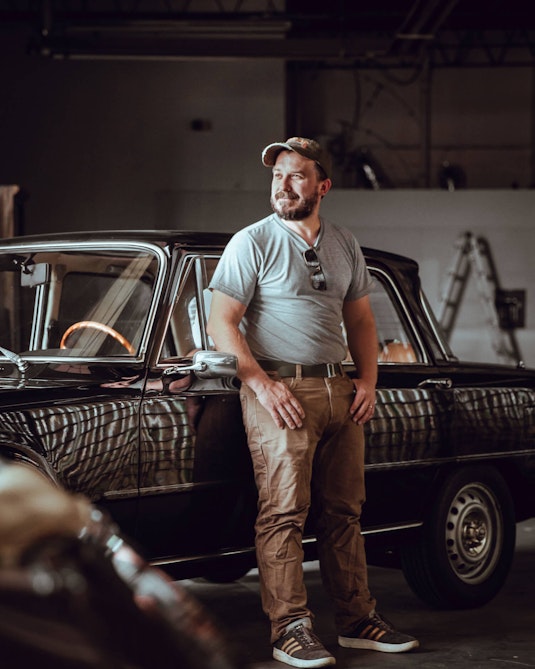 A red-haired, bearded man standing in front of a shiny collector car.