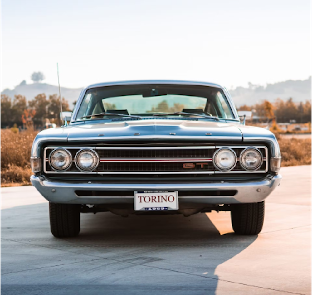 A 1969 Ford Torino faces the camera with a front license plate that reads TORINO on a road with mountains and trees in the background