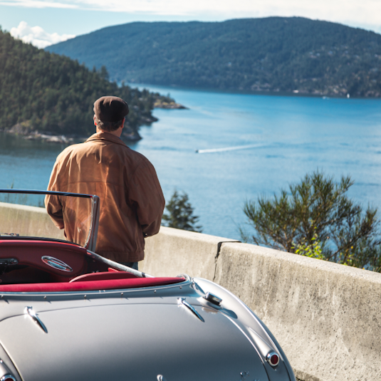 A man stands outside a silver classic car on a bridge overlooking water and mountains.