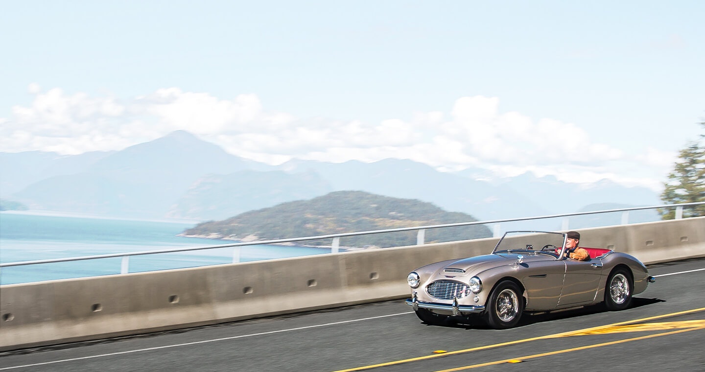 A collector convertible, driving on a freeway with mountains in the background.