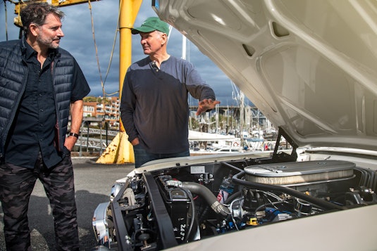 Two men in a harbor admire the engine of a white collector vehicle.