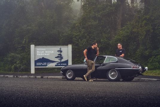 2 men stand on opposites of a black 1964 Jaguar E-Type Replica near a sign that says Entering Blue Ridge Parkway with trees in the background