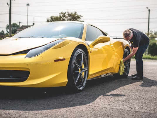 A man bends over and wipes the wheel of a pristinely cared-for yellow sports car with a cloth.