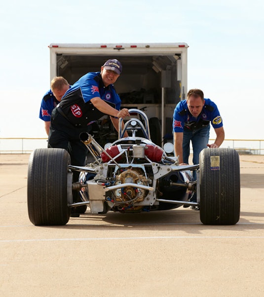 Three men pushing a collector race car, with a trailer in the background.