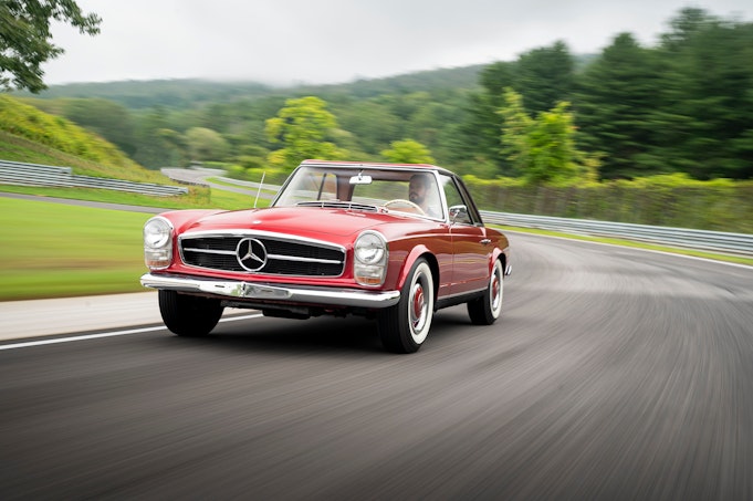 A classic red Mercedes-Benz speeds down a secluded curvy road through a lush green countryside.