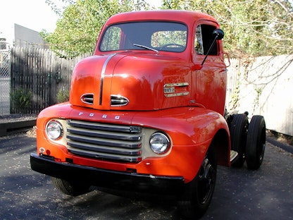 A red collector semi truck parked on concrete, with a wooden fence and trees in the background.